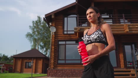 Crooked-brunette-with-inflated-press-muscles-and-black-shorts,-drinking-protein-after-a-workout-on-the-street-against-the-background-of-a-huge-house-with-large-Windows