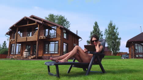 A-beautiful-brunette-in-a-swimsuit-lying-on-a-sun-lounger-near-her-chic-mansion-with-large-Windows-smiles-and-waves-her-hand-to-friends-and-works-remotely-on-a-tablet-computer