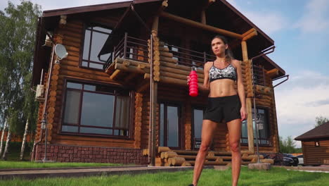 Crooked-brunette-with-inflated-press-muscles-and-black-shorts,-drinking-protein-after-a-workout-on-the-street-against-the-background-of-a-huge-house-with-large-Windows
