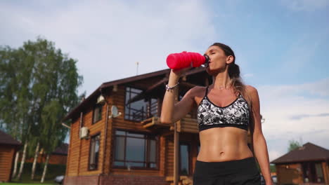 Sports-woman-with-a-beautiful-press-in-a-black-top-on-the-background-of-a-wooden-mansion-after-a-workout-drinking-water-from-a-bottle.
