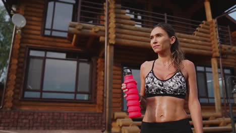 Sports-woman-with-a-beautiful-press-in-a-black-top-on-the-background-of-a-wooden-mansion-after-a-workout-drinking-water-from-a-bottle.