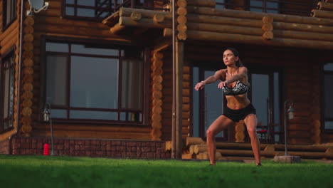 A-slender-young-woman-in-black-sportswear-performs-squats-on-the-grass-near-her-country-house