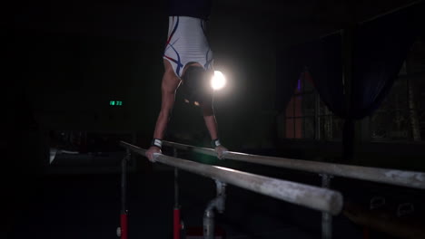 El-Gimnasta-Acróbata-Masculino-Realiza-Una-Parada-De-Manos-En-Barras-Paralelas-En-Una-Habitación-Oscura-En-Cámara-Lenta-Compartiendo-Un-Salto-Mortal-Y-Aterrizando-En-El-Suelo.-Entrenamiento-Antes-De-Los-Juegos-Olímpicos