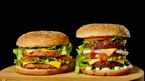 Close-up-of-two-appetizing-burgers-with-sesame-buns-rotating-on-black-background,-of-fast-food-seamless-looping-shot-.