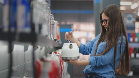 Counter-of-electronics-store.-Young-beautiful-brunette-woman-holding-an-electric-kettle-in-her-hands,-studying-the-price-tags,-characteristics-and-design-of-the-model