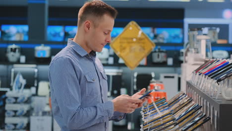 Young-handsome-man-in-blue-shirt-chooses-a-new-mobile-phone-in-an-electronics-store.-Modern-technology-purchase