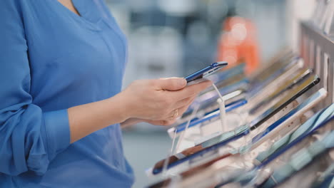 Primer-Plano-De-La-Mano-De-Una-Mujer-Que-Sostiene-Un-Nuevo-Teléfono-Inteligente-En-Sus-Manos-Y-Elige-Comprar-El-Mejor-Modelo-De-Teléfono-En-La-Tienda-De-Electrónica-Moderna