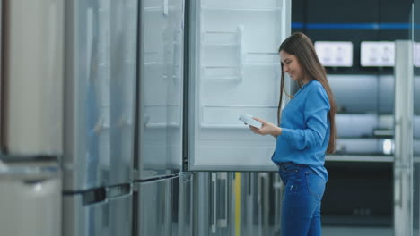 Young-sexy-brunette-woman-in-shirt-to-open-the-door-of-the-refrigerator-in-the-appliances-store-and-compare-with-other-models-to-buy-the-new-house