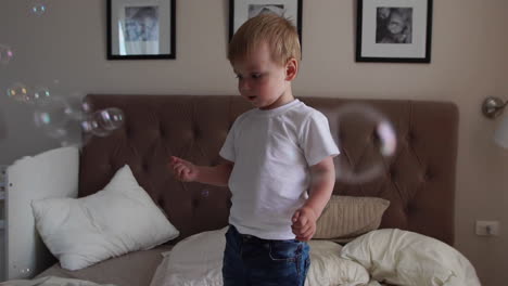 Ein-Junge-In-Einem-Weißen-T-Shirt-Und-Einer-Blauen-Jeans-Fängt-Seifenblasen-Ein,-Die-Auf-Dem-Bett-Im-Elternschlafzimmer-Stehen
