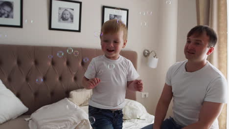 Caring-father-plays-son-looking-at-soap-bubbles,-the-boy-is-happy-jumping-on-the-bed