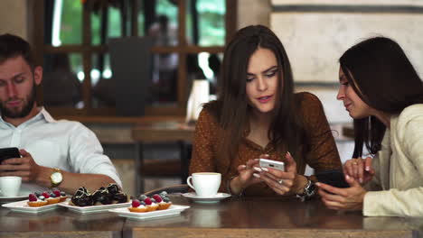 One-on-one-meeting.Two-young-business-women-sitting-at-table-in-cafe.Girl-shows-her-friend-image-on-screen-of-smartphone.-On-table-is-closed-notebook.Meeting-friends.