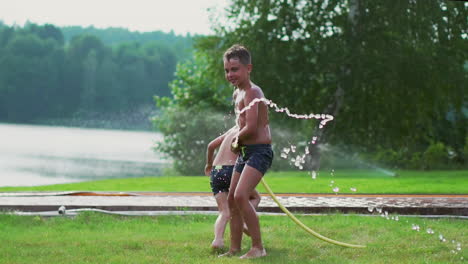 Junge-In-Sommerbadehose-Gießt-Wasser-Auf-Seinen-Jüngeren-Bruder,-Der-Sich-Im-Park-Auf-Dem-Gras-In-Der-Nähe-Des-Sees-Vergnügt