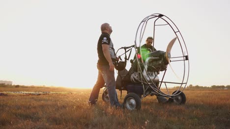 Motor-paraglider-stands-in-a-field-at-sunset-with-a-wooden-propeller,-two-pilots-warm-up-the-engine-before-the-flight.-A-test-run-of-all-systems