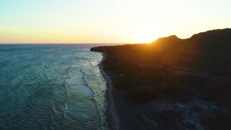 Drone-rises-high-above-rocky-caribbean-beach-shoreline-as-waves-crash-and-sun-sets-below-horizon-for-sunset