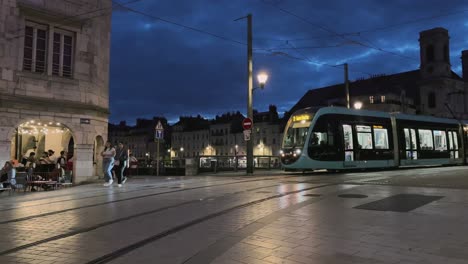 Night-street-with-tram-crossing-over-bridge-and-people-walking-in-Besancon,-France-with-streetlights-and-cafe