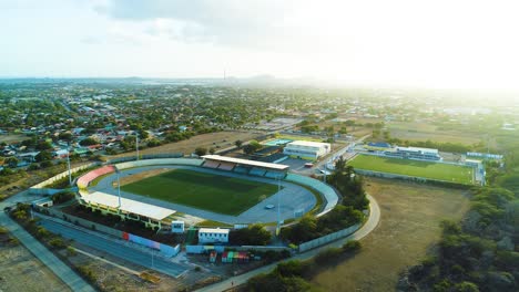 Golden-hour-light-spreads-across-SDK-Stadion-Ergilio-Hato-football-stadium-in-curacao,-aerial-establish