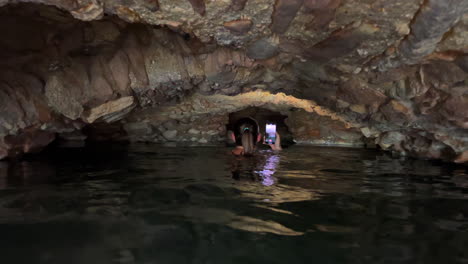 Chica-Explorando-Con-Linterna-De-Teléfono-Cuevas-Oscuras-De-Baños-Romanos-De-Casares-Manilva,-Gente-Nadando-En-Manantiales-Naturales-Curando-Agua-De-Azufre-En-España,-Buenos-Beneficios-Para-La-Salud,-Toma-De-4k