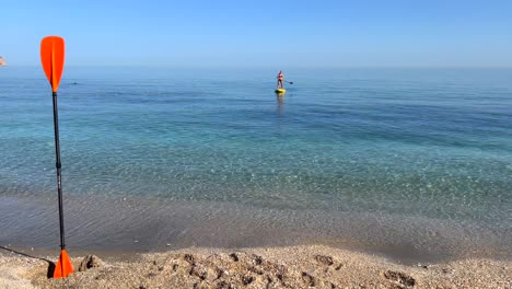 Girl-puddle-surfing-in-the-sea-in-Nerja-Malaga,-stand-up-puddle-boarding-on-a-sunny-day-in-Spain,-fun-vacation-activity,-adventurous-water-sport,-4K-shot