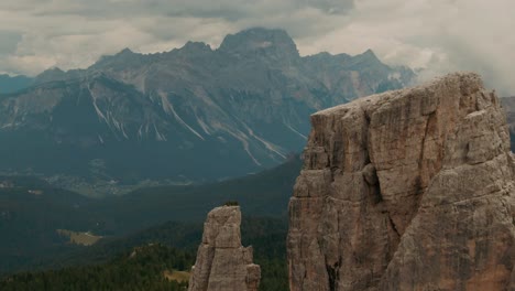 Aerial-view-flying-past-massive-rocks-to-high-mountains-in-the-background,-green-forest-at-the-bottom,-cloudy-day,-cinematic-color-grade