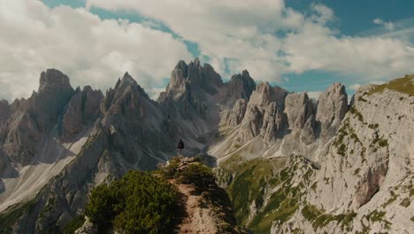 Parralax-drone-shot-with-lone-tourist-watching-panoramic-view-with-tall-steep-rocky-mountains,-partly-clouded-sky-in-the-background,-hiking-in-the-Alps,-majestic-landscape,-cinematic-color-grade
