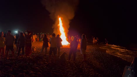 Gente-Parada-Alrededor-De-Un-Tradicional-Festival-De-Verano-Con-Hogueras-En-La-Playa-En-La-Celebración-De-San-Juan-En-Marbella,-España,-Disfrutando-De-Una-Fiesta-Divertida,-Un-Gran-Fuego-Ardiente-Y-Llamas-Calientes-Por-La-Noche,-Toma-De-4k