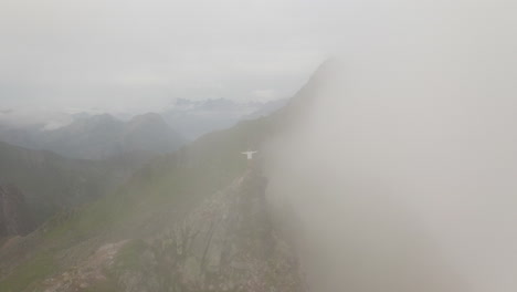Revealing-aerial-around-young-hiker-man-standing-on-Mountain-Peak-Summit,-moving-through-the-clouds-Fløya-and-Djevelporten-above-Svolvær-in-Lofoten,-Norway,-aerial-footage