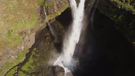 Aerial-drone-shot-of-a-beautiful-waterfall-in-Iceland-on-a-sunny-day-with-birds-flying-and-a-small-rainbow-In-front-of-the-mossy-green-cliffs-and-rocks
