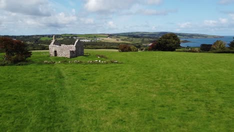 Aerial-view-of-Capel-Lligwy-ruined-chapel-on-Anglesey-island-coastline-descending-to-grassy-field,-North-Wales