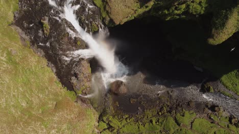 Top-down-aerial-drone-shot-of-a-beautiful-waterfall-in-Iceland-on-a-sunny-day-with-birds-flying-and-a-small-rainbow-in-front-of-the-mossy-green-cliffs-and-rocks