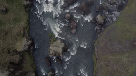 Top-down-aerial-drone-shot-of-a-beautiful-river-rapids-in-Iceland-on-a-dark-foggy-day-in-front-of-the-mossy-green-cliffs-and-rocks