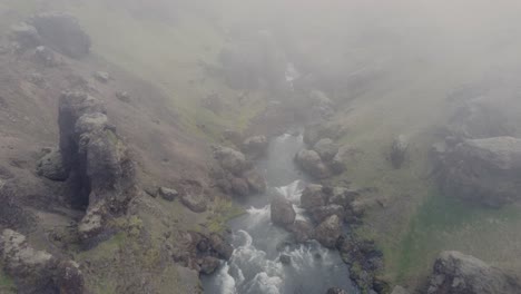 Stimmungsvolle-Luftdrohnenaufnahme-Eines-Wunderschönen-Flusses-Und-Wasserfalls-In-Island-An-Einem-Dunklen,-Nebligen-Tag-Vor-Den-Moosgrünen-Klippen-Und-Felsen