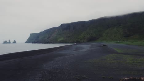 Orbiting-aerial-drone-shot-of-beautiful-blue-waves-crashing-on-a-black-sand-beach-in-Iceland-with-a-group-of-Icelandic-horses-and-huge-rock-formations-in-the-distance
