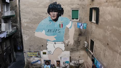 Aerial-tilt-up-shot-of-Diego-Maradona-mural-on-wall-of-old-block-in-city-of-Naples,-Italian-legend