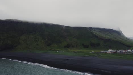 Toma-Aérea-De-Drones-De-Hermosas-Olas-Azules-Rompiendo-En-Una-Playa-De-Arena-Negra-En-Islandia-Y-Enormes-Formaciones-Rocosas-En-La-Distancia