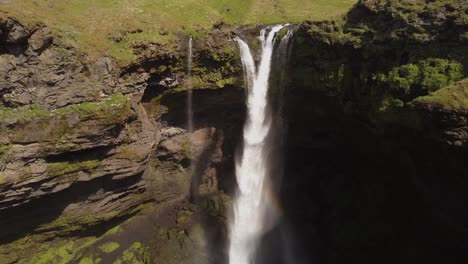 Aerial-drone-shot-of-a-beautiful-waterfall-in-Iceland-on-a-sunny-day-with-birds-flying-and-a-small-rainbow