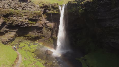 Wide-angle-receding-aerial-drone-shot-of-a-beautiful-waterfall-in-Iceland-on-a-sunny-day-with-birds-flying-and-a-small-rainbow-in-front-of-the-mossy-green-cliffs-and-rocks