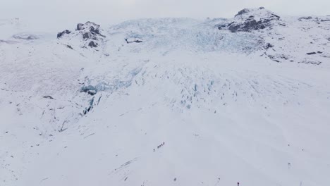Aerial-landscape-view-over-people-hiking-towards-ice-formations-in-Falljokull-glacier-covered-in-snow,-Iceland