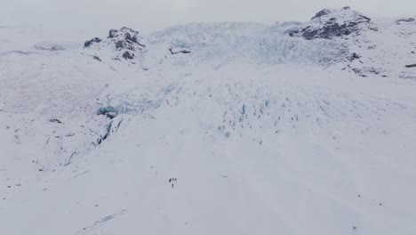 Aerial-view-over-people-hiking-towards-ice-formations-in-Falljokull-glacier-covered-in-snow,-Iceland