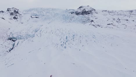 Aerial-panoramic-view-over-people-hiking-towards-ice-formations-in-Falljokull-glacier-covered-in-snow,-Iceland