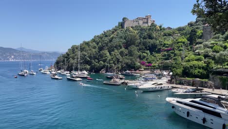 Toma-Panorámica-De-Vistas-Panorámicas-De-Casas-Coloridas,-Castello-Brown,-Un-Castillo-Histórico-Encaramado-En-Una-Colina-Y-Barcos-Navegando-Y-Amarrando-En-El-Pequeño-Puerto-De-Portofino,-Italia