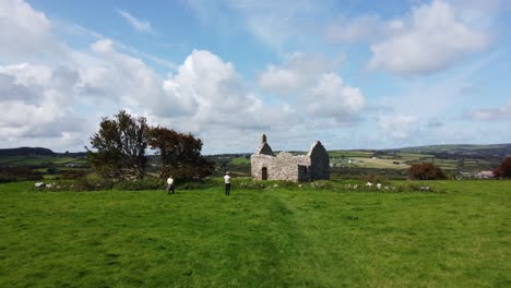 Aerial-view-hipster-tourists-sightseeing-Capel-Lligwy-ruined-chapel-on-Anglesey-island-coastline,-North-Wales