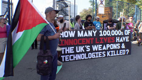 Protestors-hold-a-large-black-banner-that-reads,-“How-many-1000,000s-of-innocent-lives-have-the-UK’s-weapons-and-technologies-killed