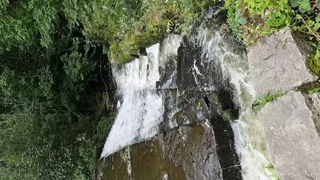 VERTICAL-slow-motion-water-cascading-down-old-Welsh-industrial-mining-stream-reservoir-towards-woodland-forest