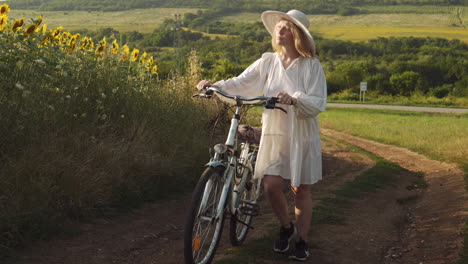 Girl-in-white-dress-pushes-bike-in-rural-sunflower-scene-at-golden-hour-slow-mo