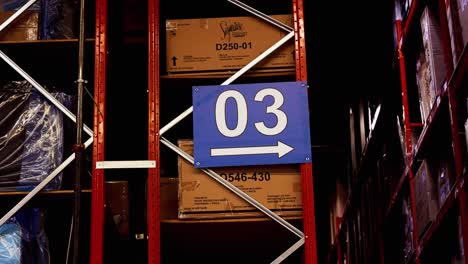 Ein-Großes-Schild-Mit-Der-Aufschrift-„03-Warehouse-Logistics-Distribution-Center“-Mit-Einem-Pfeil-Zeigt-Direkt-Nach-Unten-Auf-Eine-Reihe-Von-Lagerregalen-In-Der-Fabrik,-In-Denen-Sich-Die-Für-Ashley-Typischen-Möbelkisten-Mit-Waren-Und-Produkten-Befinden