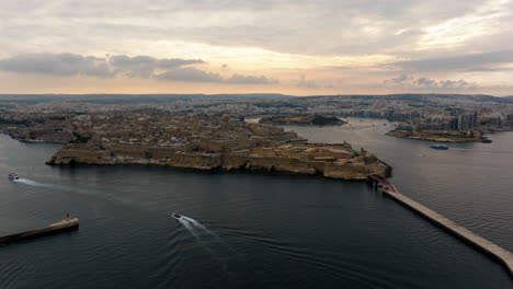 Aerial-view-following-a-boat-in-front-of-the-Valletta-cityscape,-cloudy-sunset-in-Malta