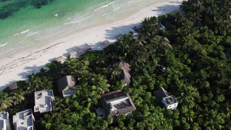 Aerial-Orbit-top-down-shot-of-huts-and-cabins-surrounded-by-jungle-and-palm-trees-in-front-of-a-white-sandy-beach-with-crystal-clear-water-in-Tulum,-Mexico