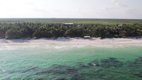 Aerial-Push-in-shot-as-crystal-clear-ocean-waves-roll-through-in-a-white-sand-beach-with-palm-trees-and-huts-and-cabins-in-Tulum,-Mexico