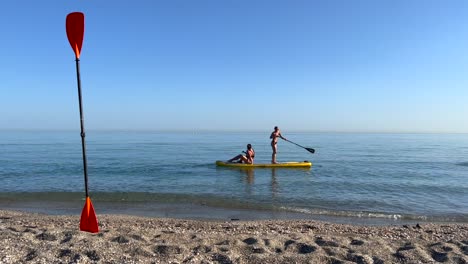 Two-girls-puddle-surfing-in-the-sea-in-Nerja-Malaga,-stand-up-puddle-boarding-on-a-sunny-day-in-Spain,-fun-vacation-activity,-adventurous-water-sport,-4K-shot