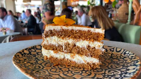 Delicious-carrot-cake-with-a-yellow-flower-on-a-plate-in-a-pretty-restaurant,-sweet-dessert,-unrecognizable-blurry-people-in-the-background,-4K-shot